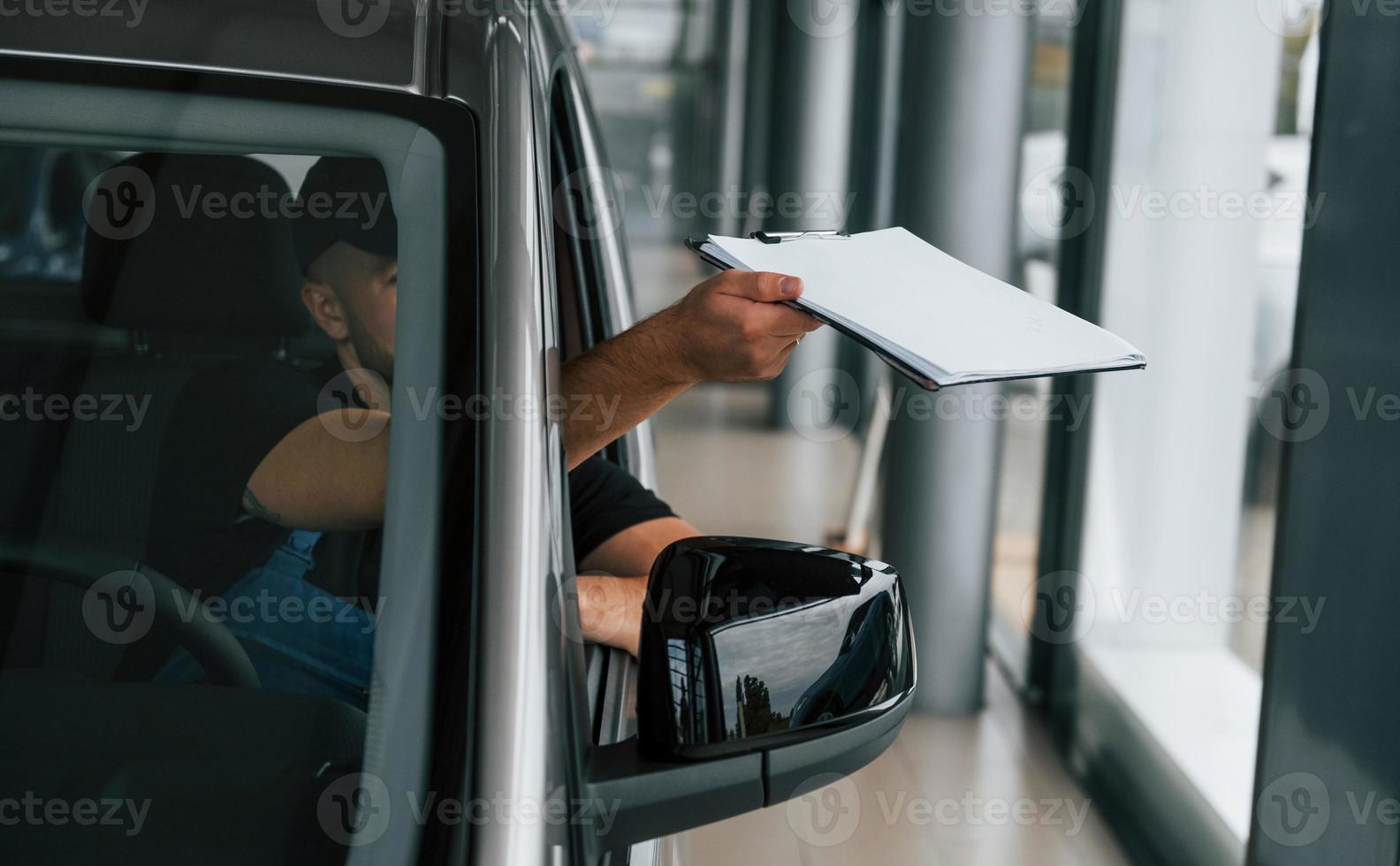 Sitting in vehicle and holding document. Delivery man in uniform is indoors with car and with order photo