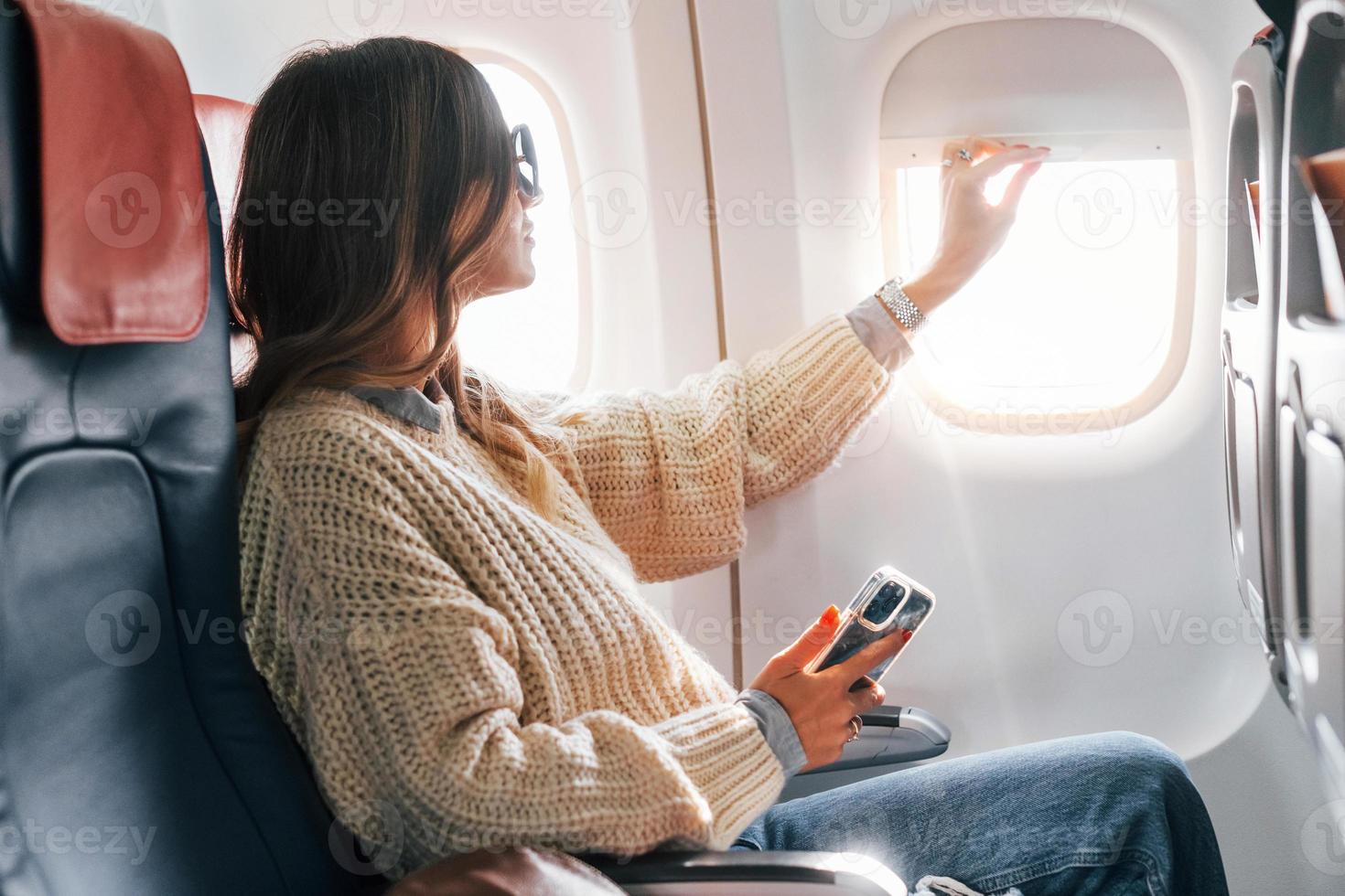 Sitting with phone in hands. Young female passanger in casual clothes is in the plane photo