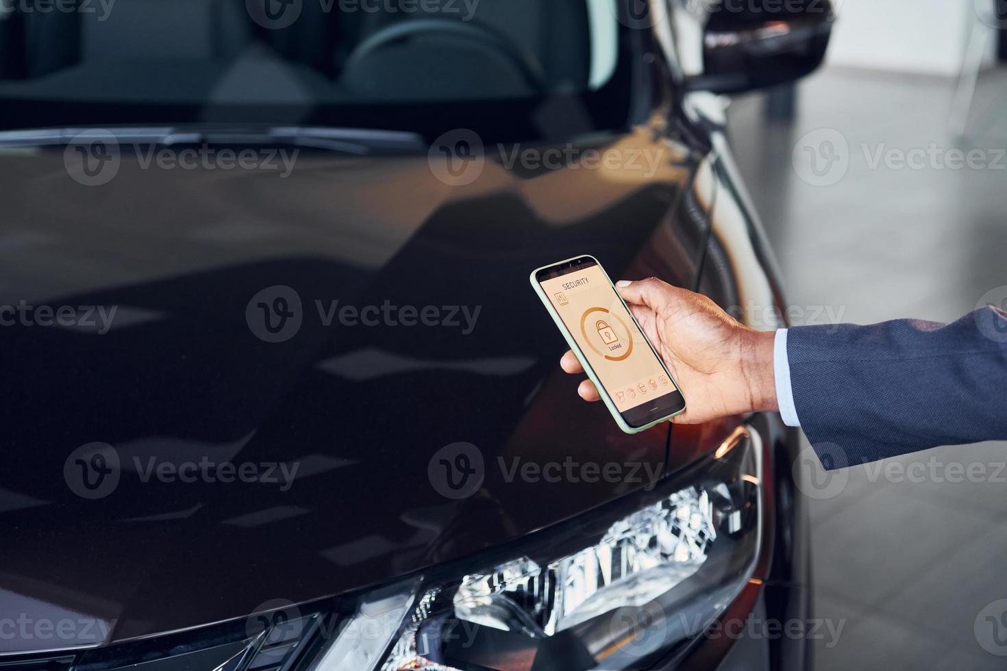 Using smartphone to open door. Young african american businessman in black suit is the autosalon photo