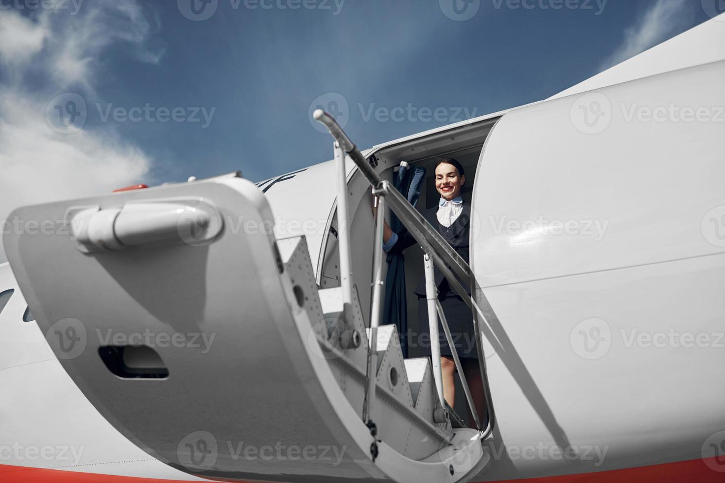 At daytime. Young stewardess that is in formal black clothes is standing outdoors near plane photo