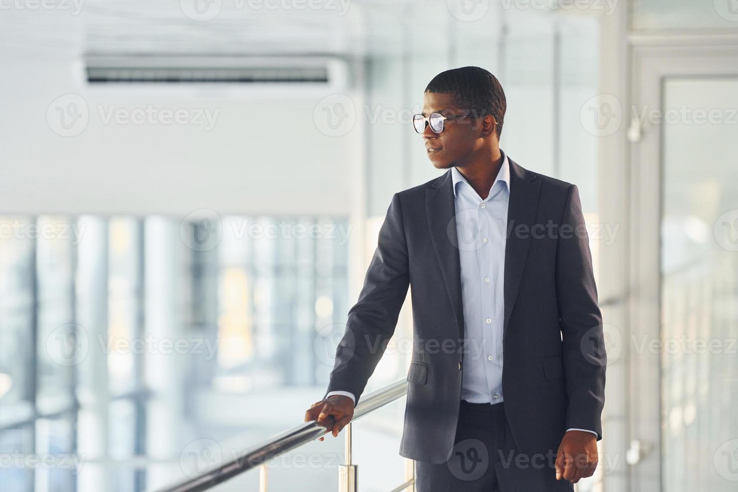 Leaning on the railings. Young african american businessman in black suit is indoors photo