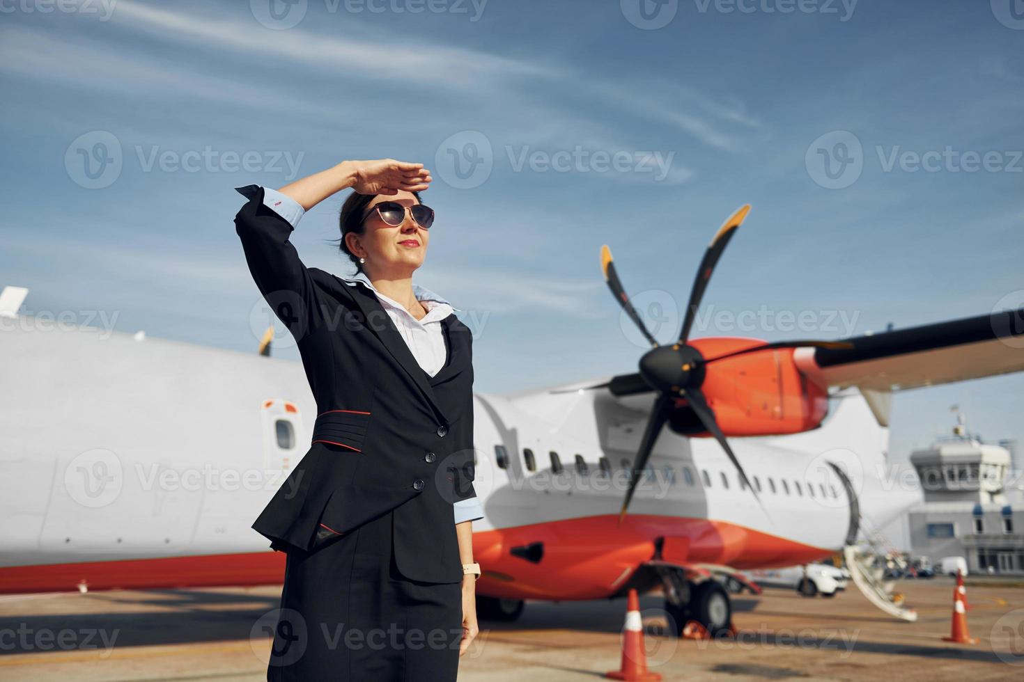 At daytime. Young stewardess that is in formal black clothes is standing outdoors near plane photo