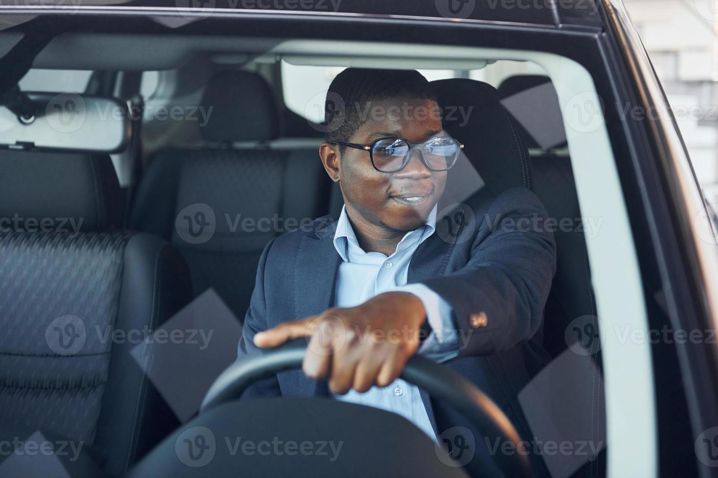 Front view. Young african american businessman in black suit is in the automobile photo