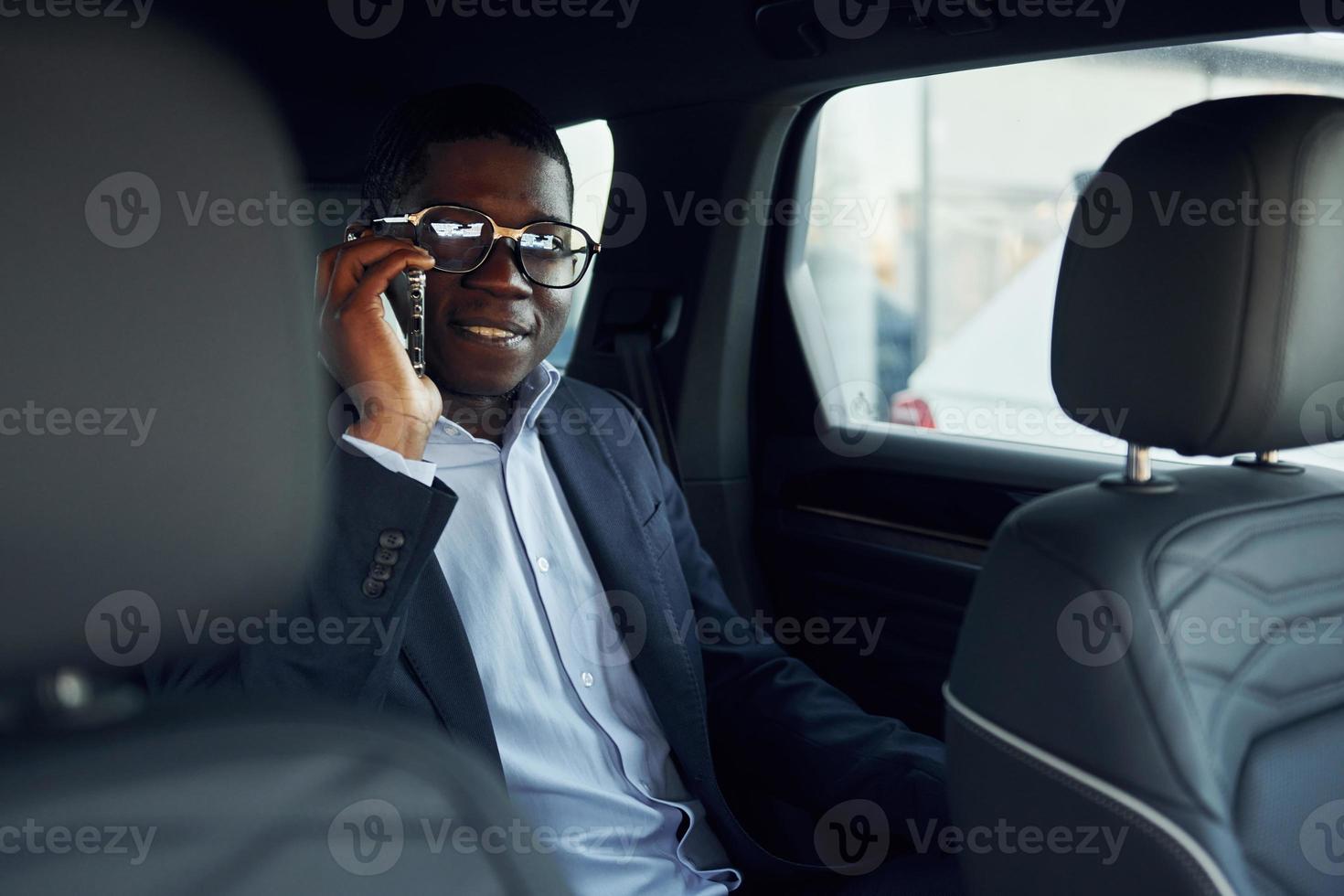 In glasses. Young african american businessman in black suit is in the automobile photo