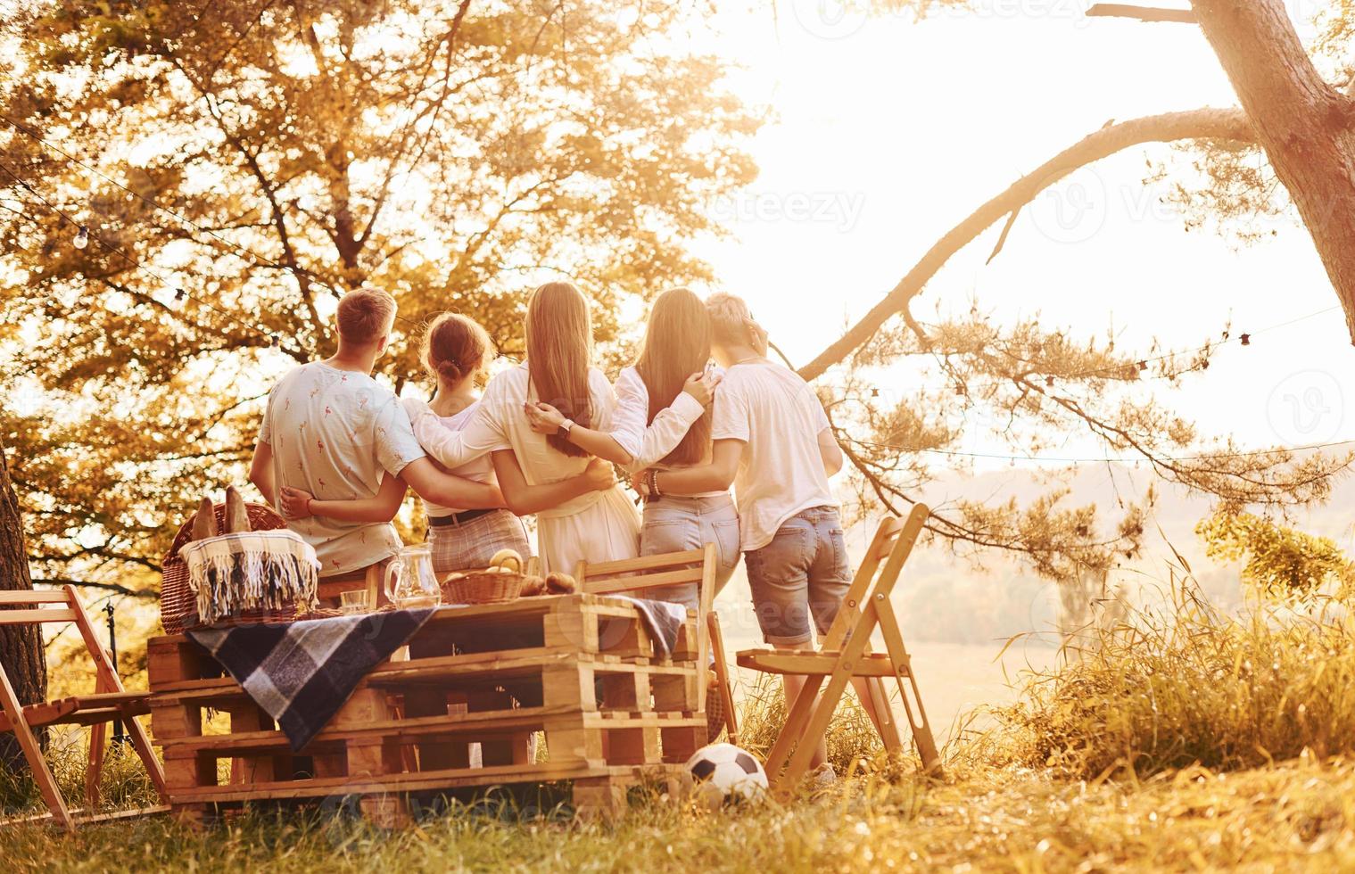 Embracing each other and looking at sunset. Group of young people have vacation outdoors in the forest. Conception of weekend and friendship photo