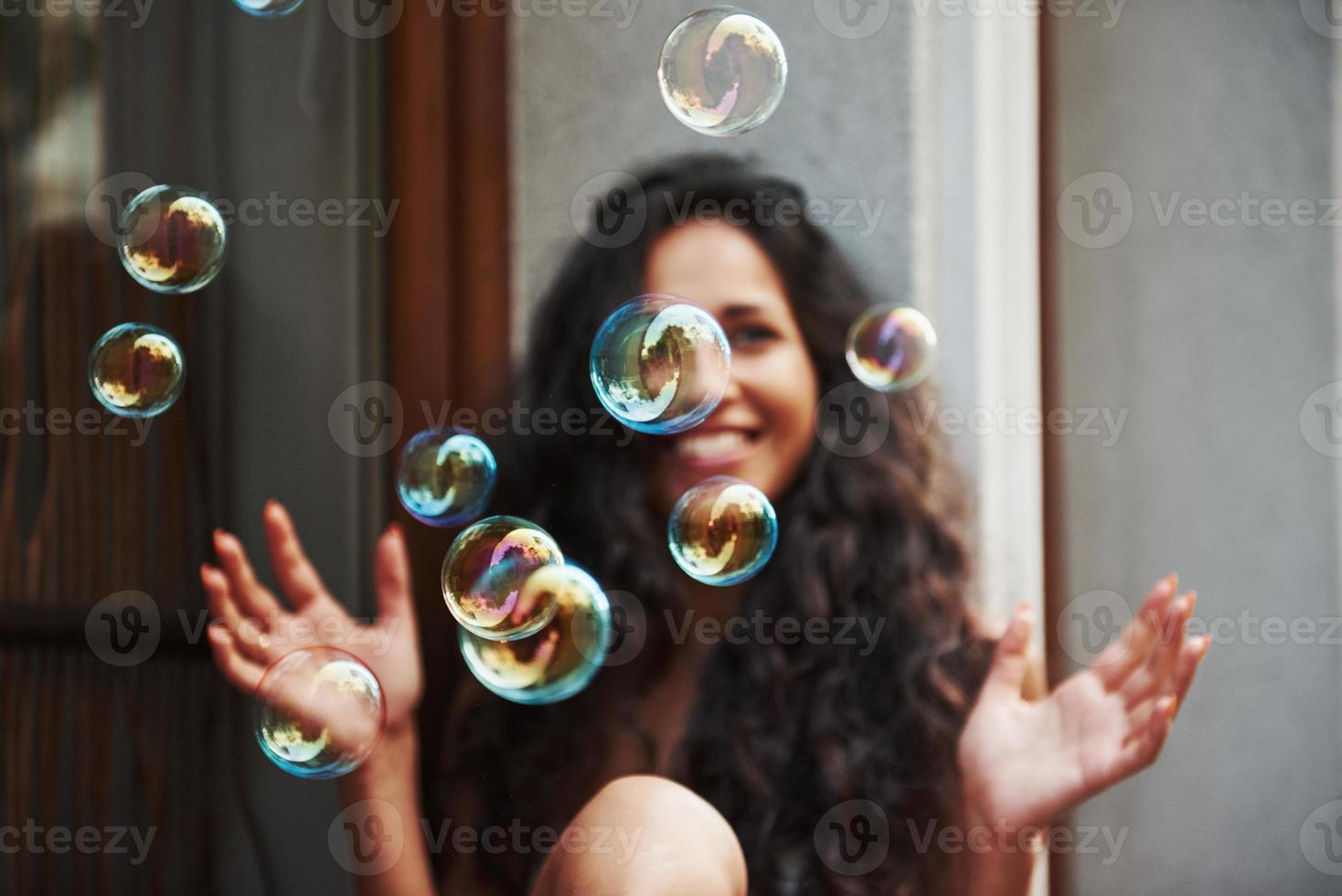 linda chica juega con burbujas como un niño. hermosa mujer con cabello negro rizado pasar un buen rato en la ciudad durante el día foto