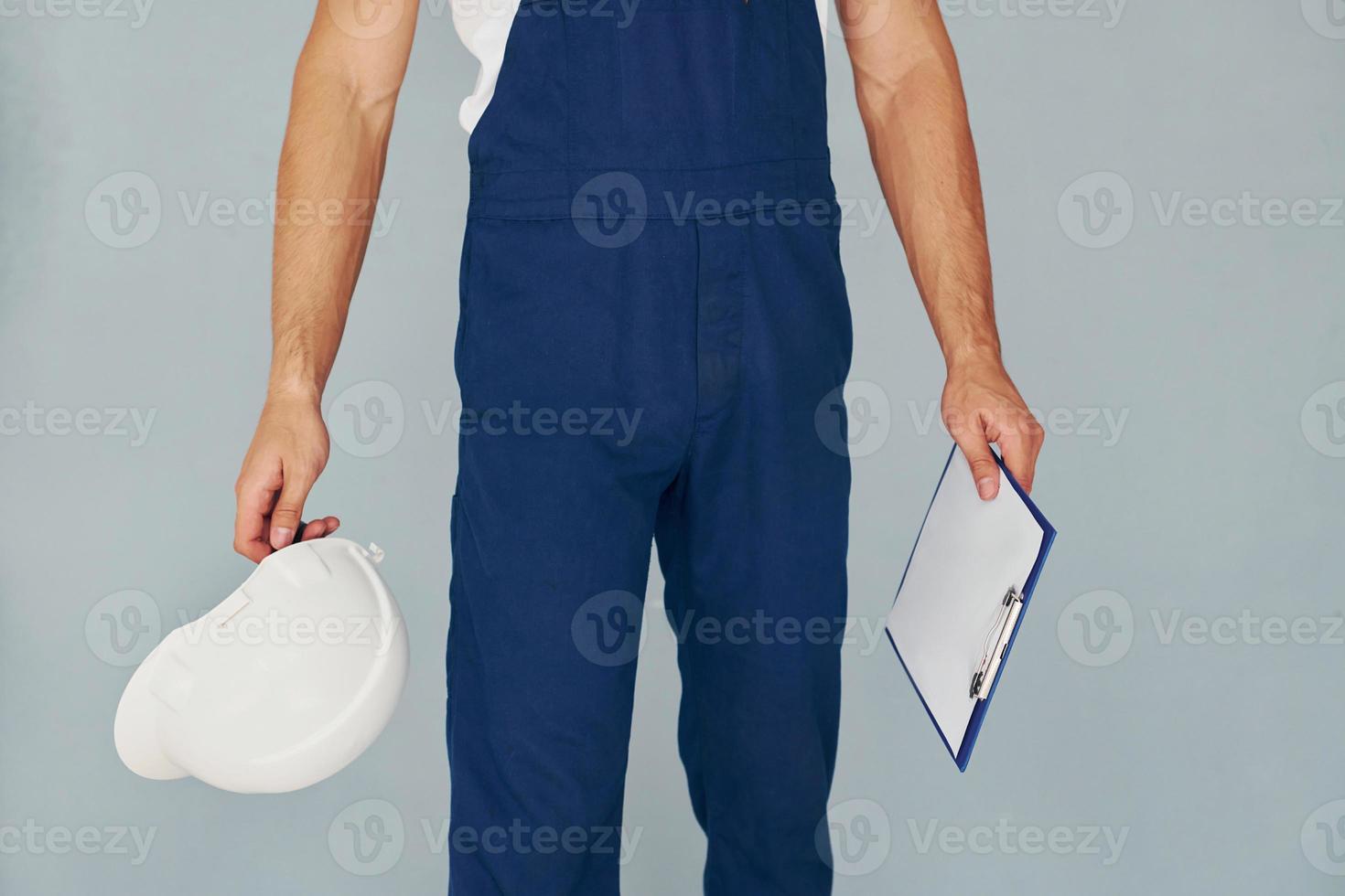 With notepad and hard hat. Male worker in blue uniform standing inside of studio against white background photo