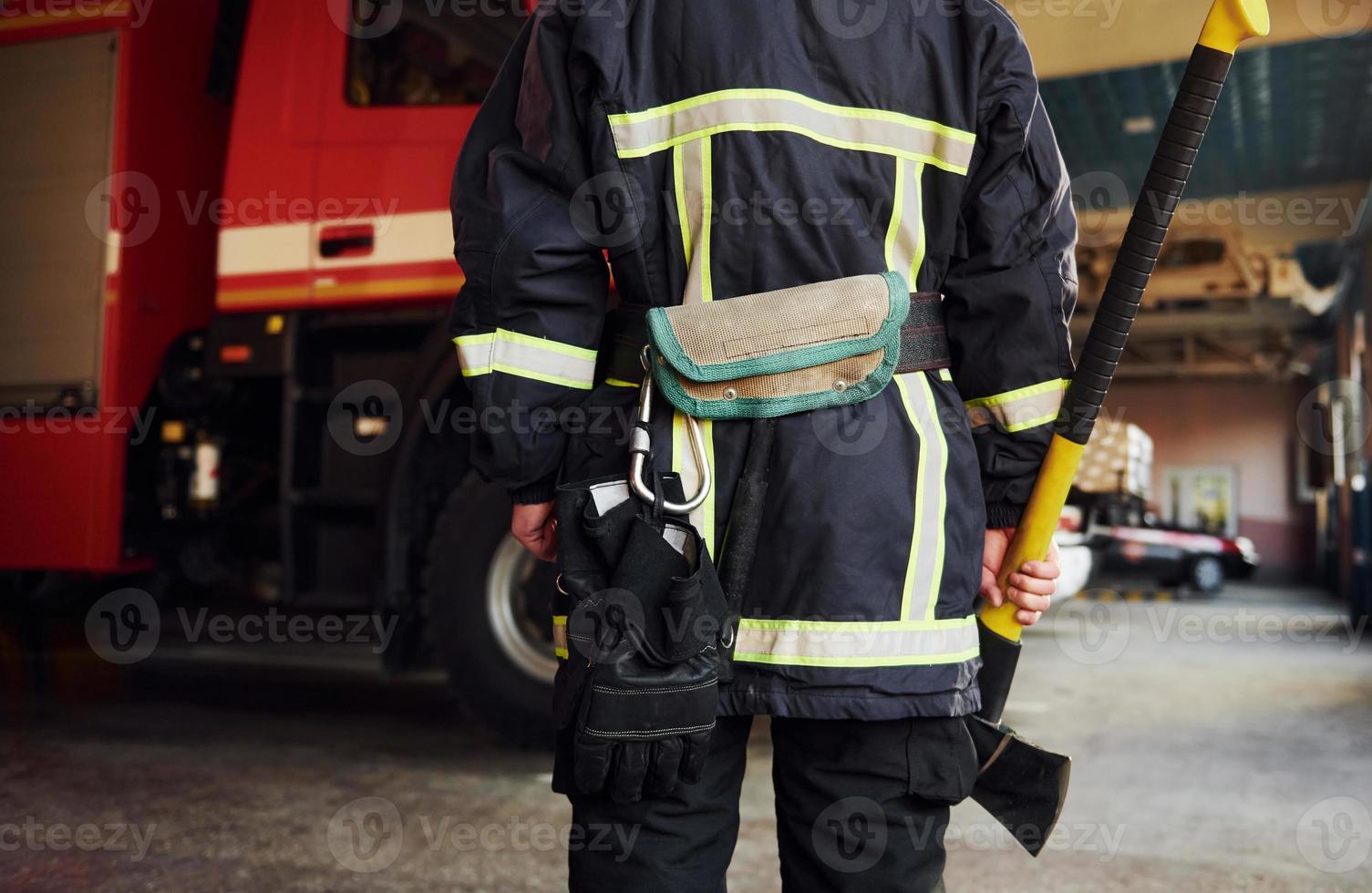 Holds axe in hands. Male firefighter in protective uniform standing near truck photo