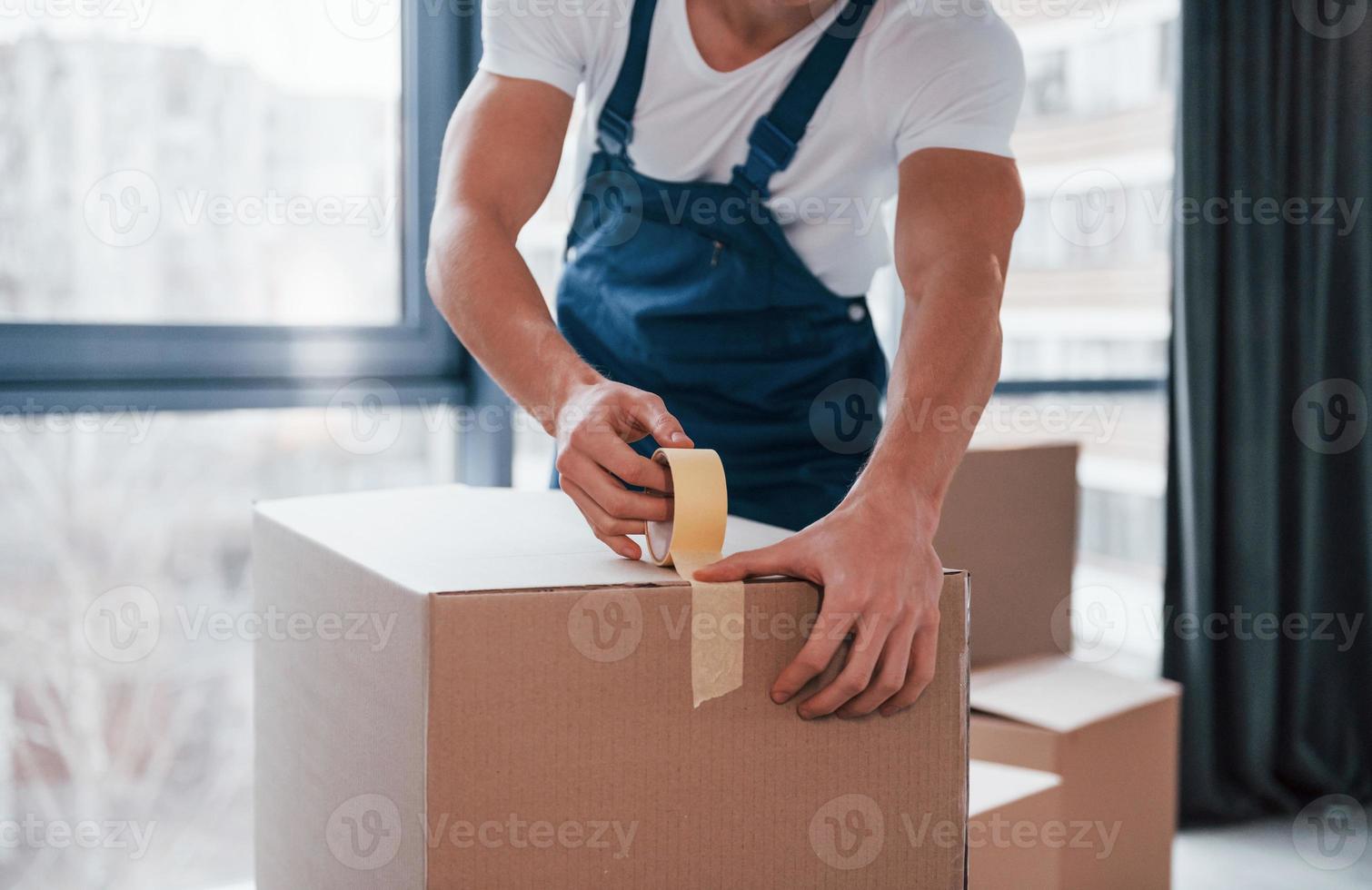 Packaging the box. Young male mover in blue uniform works indoors in the room photo