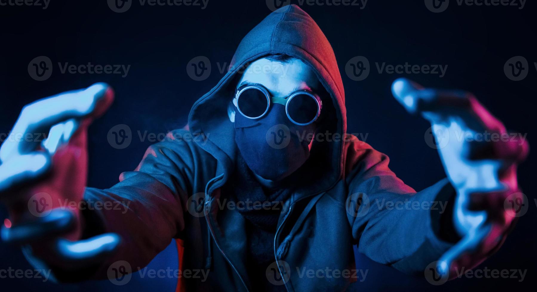 Scary guy in eyewear, mask and hood. Neon lighting. Young european man is in the dark studio photo