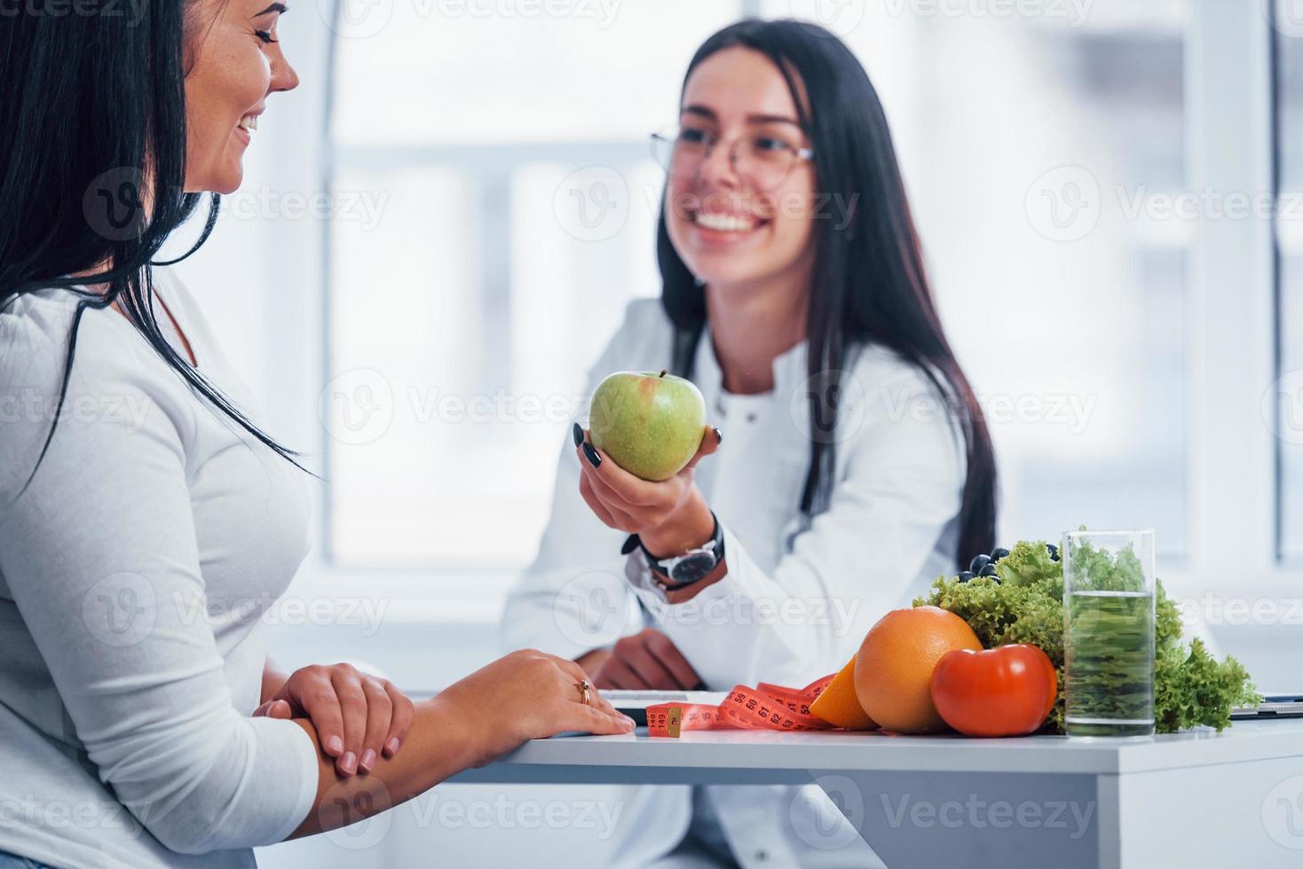 Female nutritionist holding green apple and gives consultation to patient indoors in the office photo