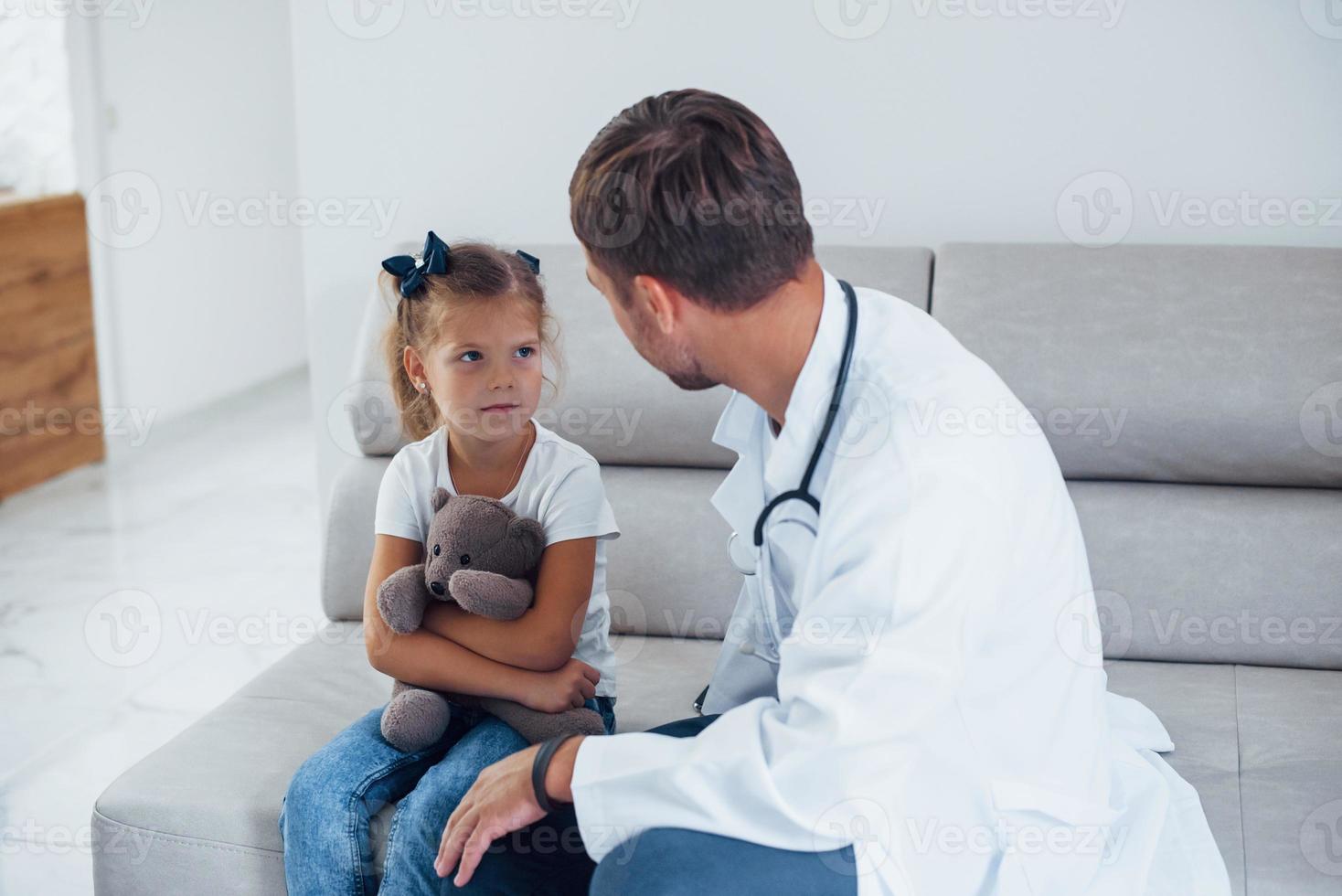 Male doctor in white uniform sits in the clinic with little girl photo