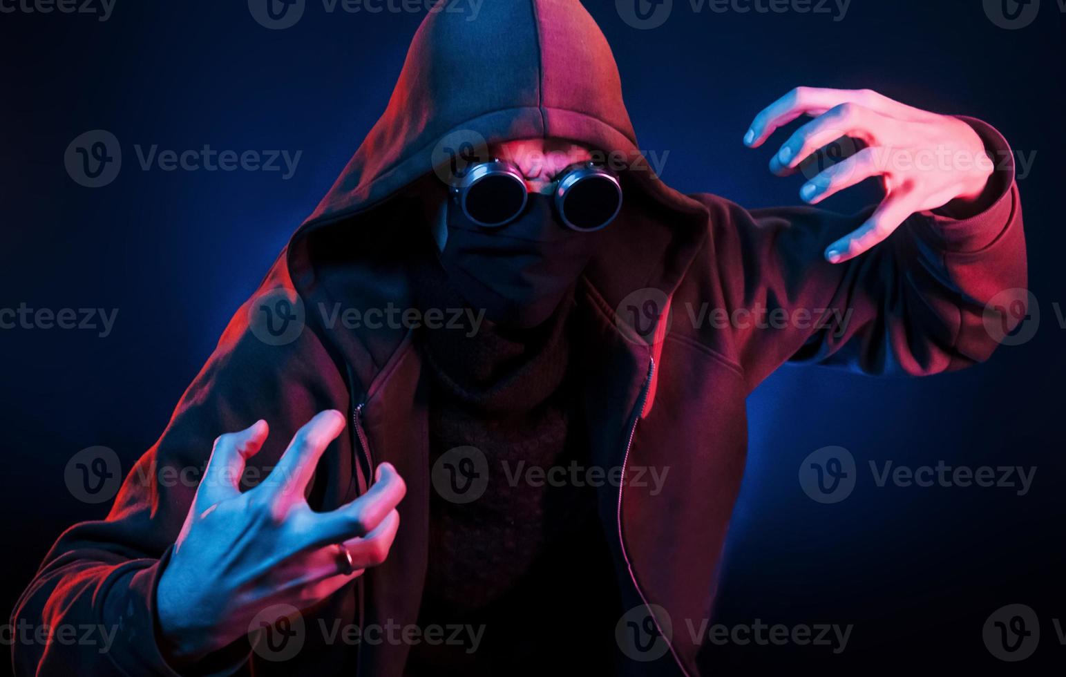 Scary guy in eyewear, mask and hood. Neon lighting. Young european man is in the dark studio photo