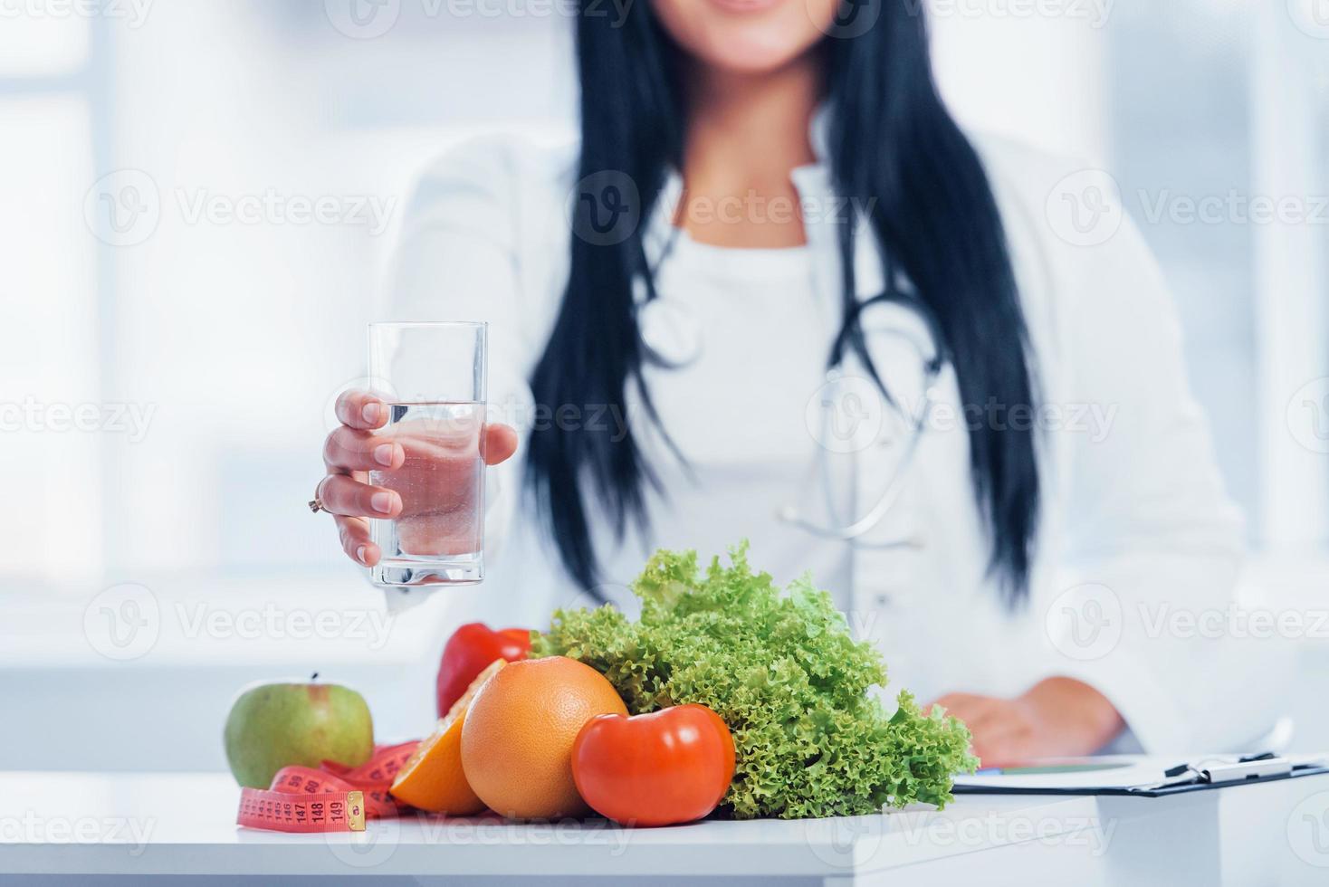 Female nutritionist in white coat sitting indoors in the office at workplace with glass of drink photo