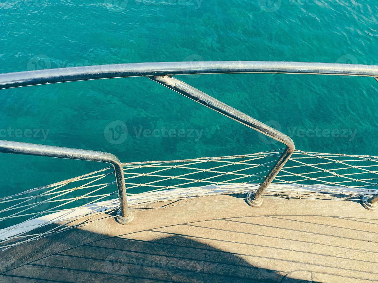 yate en mar abierto. mar con una playa de arena con el telón de fondo de una montaña. el yate tiene un tablero de madera con pasamanos de metal foto