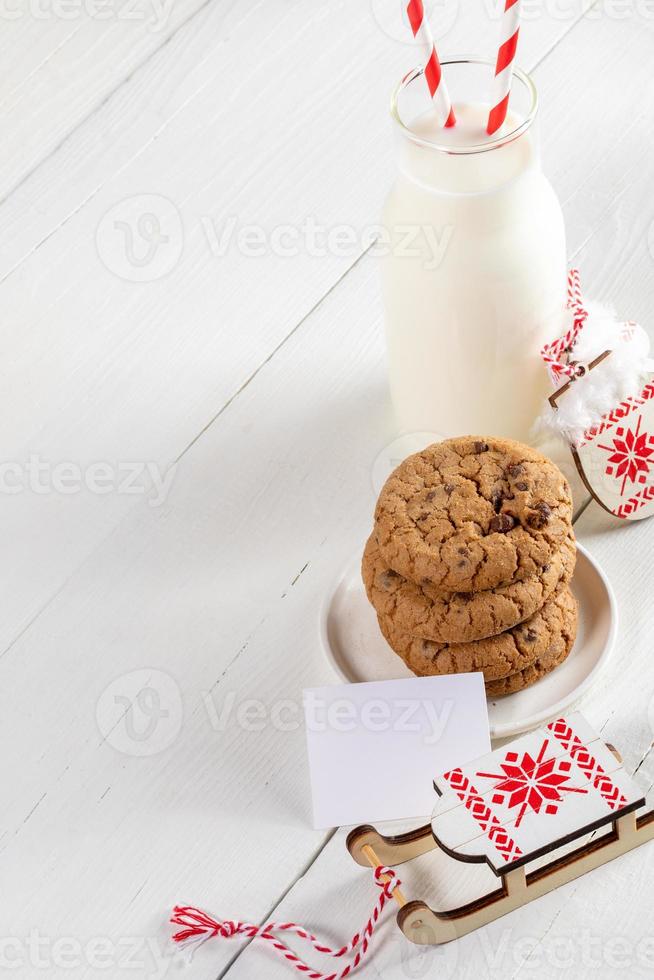 botella de leche, galletas, nota en blanco, guantes decorativos, trineo sobre tablas diagonales de madera blanca. Navidad. foto