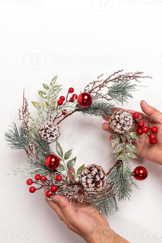 Woman hands are holding handmade reusable Christmas wreath made of tree branches, red berries, cones on white backdrop. photo