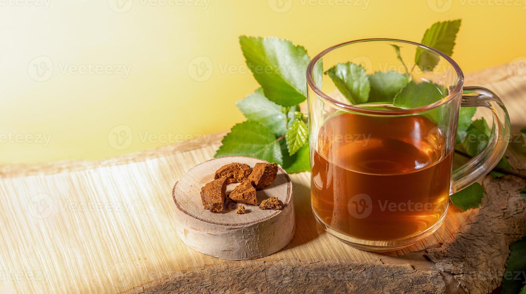 taza de vidrio de bebida curativa de chaga de hongos de abedul, trozos de hongos, ramita sobre tablón de madera sobre fondo amarillo. foto