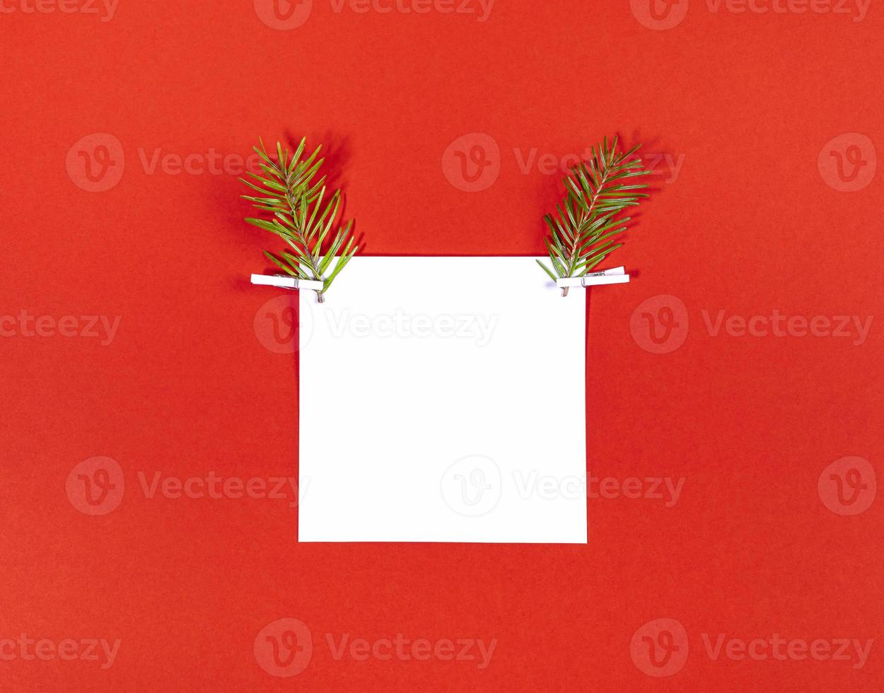 Creative Christmas deer face made of white square sheet and two fir twigs as reindeer antlers on red photo