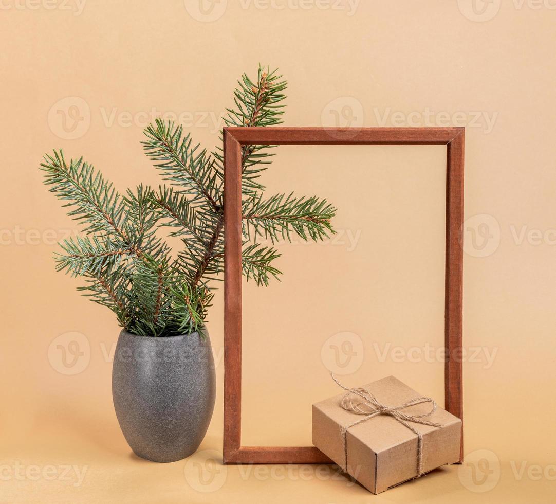 Festive trendy minimal composition with fir tree branches in grey vase, empty wooden frame, craft gift box on beige. photo