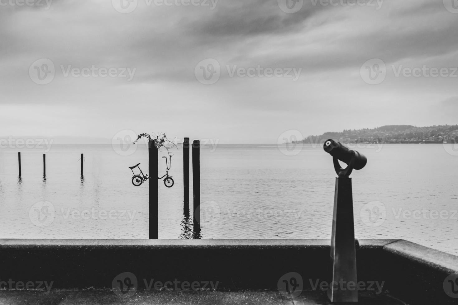 Black and white minimalist scene of a harbor with a coin-op viewer and several pillars with a bicycle in a pole photo