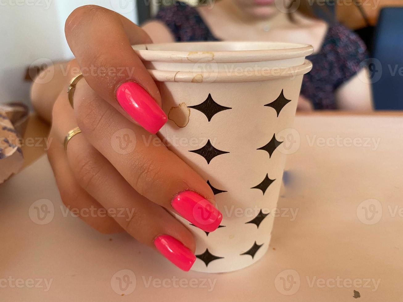 Hot drink in a craft plastic cup with a white cap in the hands of a young girl with a red manicure in a black coat close-up on the street in the fall afternoon photo