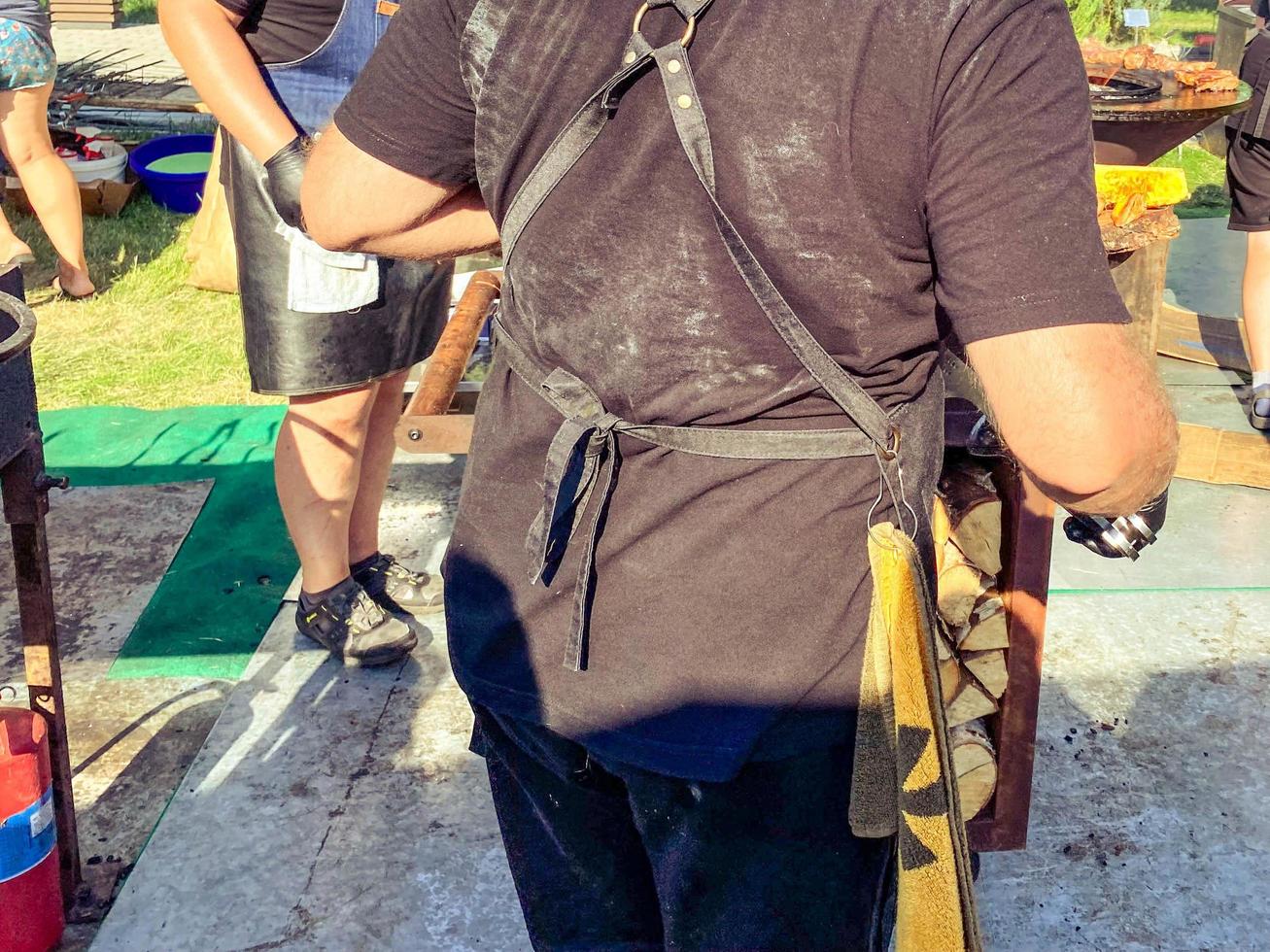 un cocinero con un delantal de cuero prepara platos a la parrilla. cocinero masculino hornea carne. plato delicioso y abundante en el festival de comida en el parque de la ciudad foto