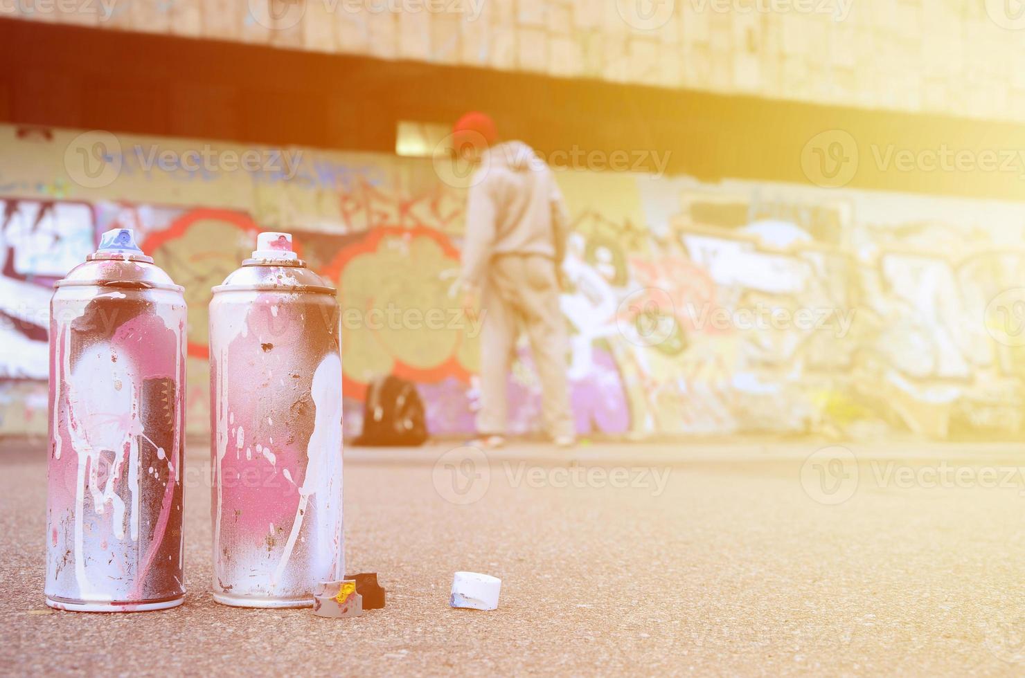 Several used spray cans with pink and white paint lie on the asphalt against the standing guy in front of a painted wall in colored graffiti drawings photo