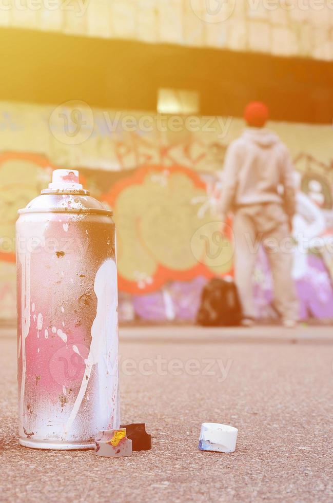 Used aerosol paint spray can with pink and white paint lie on the asphalt against the standing guy in front of a painted wall in graffiti drawings photo