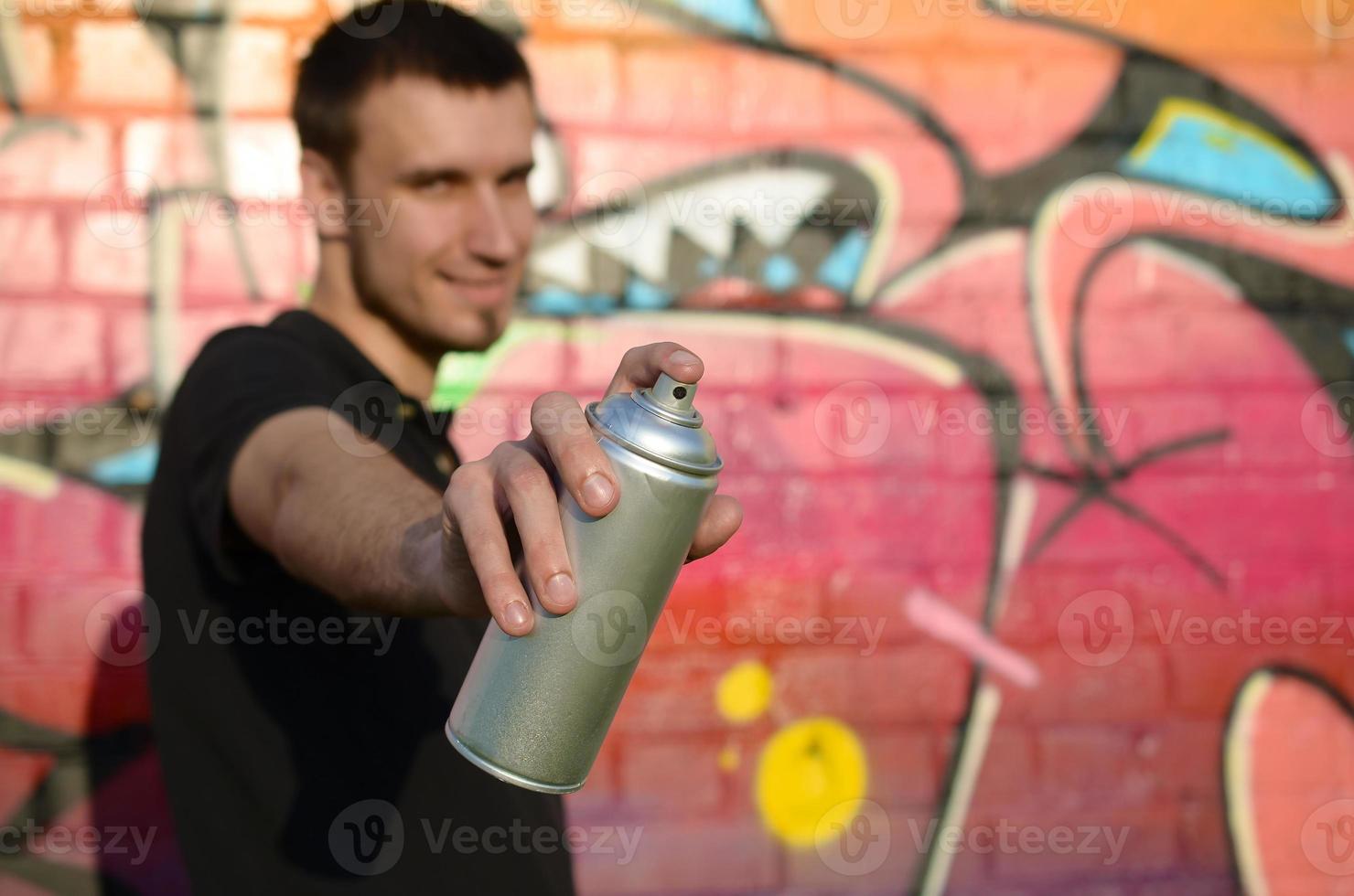 Young graffiti artist aims his spray can on background of colorful graffiti in pink tones on brick wall. Street art and contemporary painting process photo