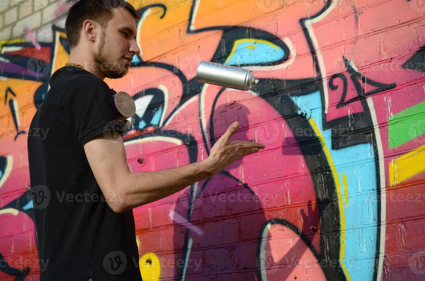 Young graffiti artist with gas mask on his neck throw his spray can against colorful pink graffiti on brick wall. Street art and contemporary painting process photo