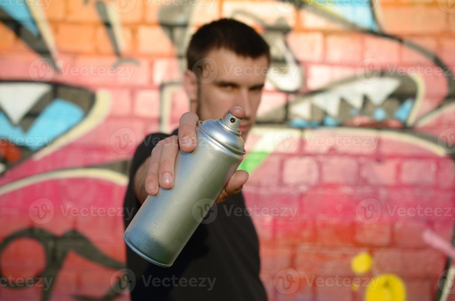 Young graffiti artist aims his spray can on background of colorful graffiti in pink tones on brick wall. Street art and contemporary painting process photo