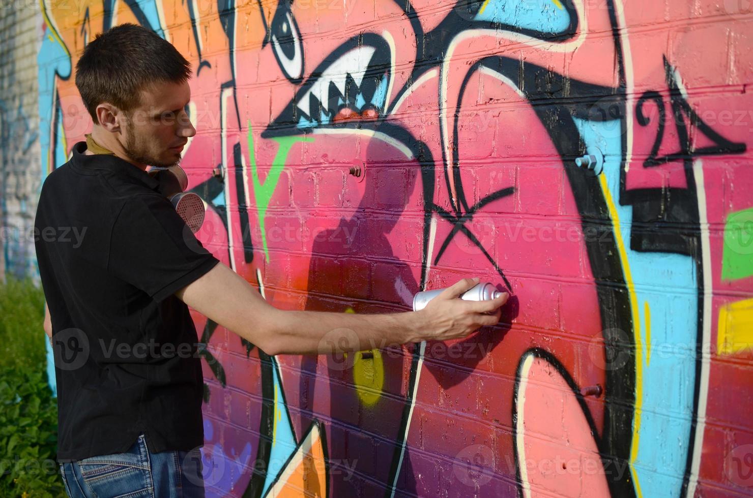 Young graffiti artist with backpack and gas mask on his neck paints colorful graffiti in pink tones on brick wall. Street art and contemporary painting process photo