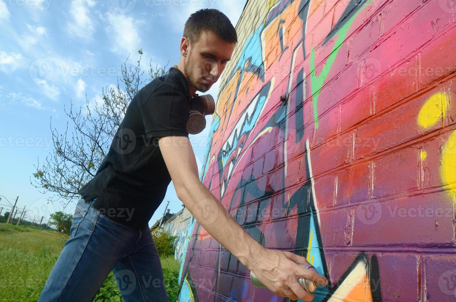 Young graffiti artist with backpack and gas mask on his neck paints colorful graffiti in pink tones on brick wall. Street art and contemporary painting process photo