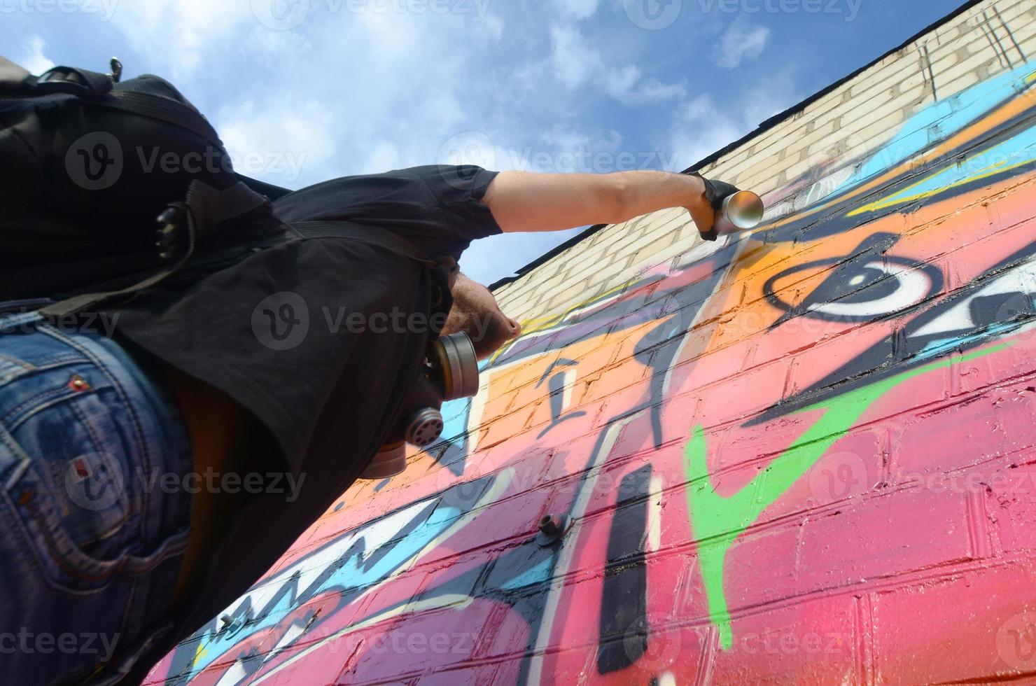 Young graffiti artist with backpack and gas mask on his neck paints colorful graffiti in pink tones on brick wall. Street art and contemporary painting process photo