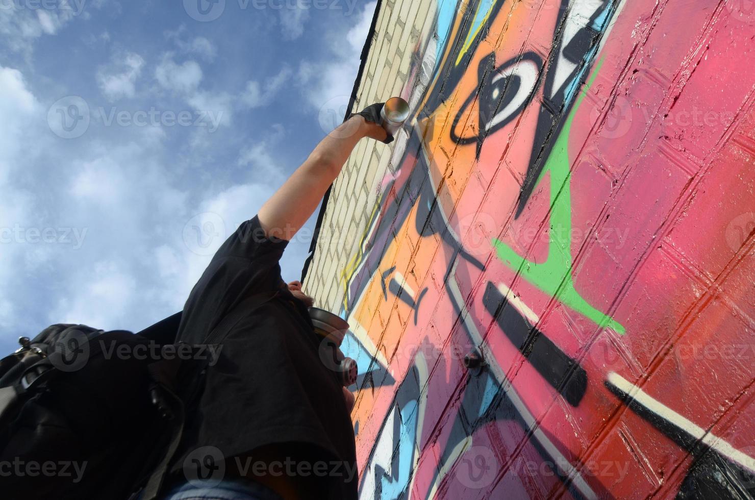 joven artista de graffiti con mochila y máscara de gas en el cuello pinta graffiti colorido en tonos rosas en la pared de ladrillo. proceso de arte callejero y pintura contemporánea foto