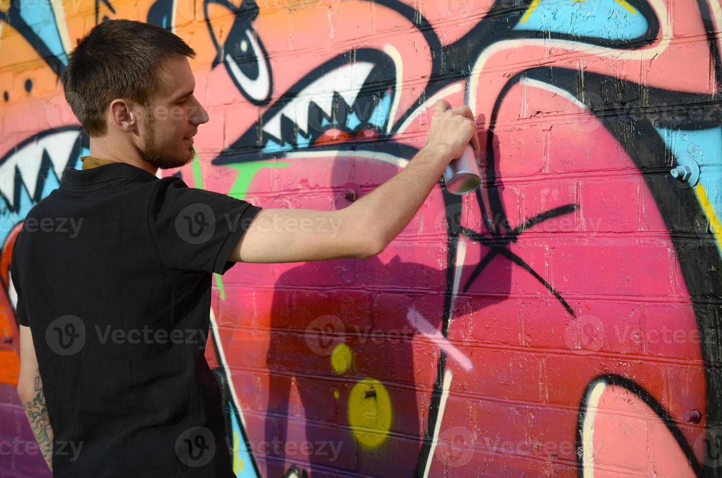 Young graffiti artist with backpack and gas mask on his neck paints colorful graffiti in pink tones on brick wall. Street art and contemporary painting process photo