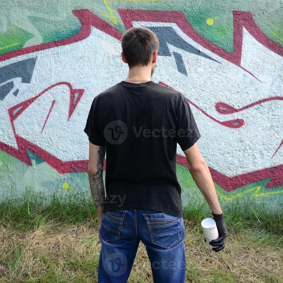 A young hooligan with a spray can stands against a concrete wall with graffiti paintings. Illegal vandalism concept. Street art photo