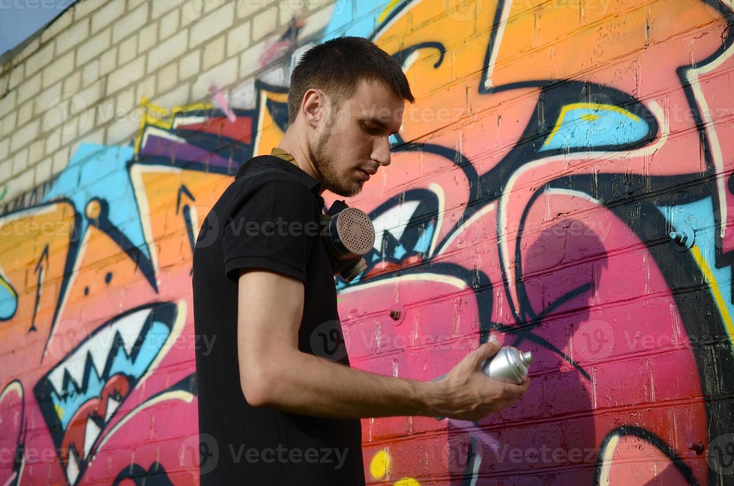 Young graffiti artist with backpack and gas mask on his neck paints colorful graffiti in pink tones on brick wall. Street art and contemporary painting process photo