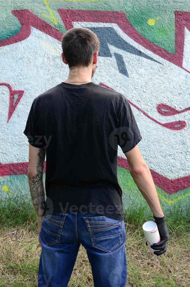 A young hooligan with a spray can stands against a concrete wall with graffiti paintings. Illegal vandalism concept. Street art photo