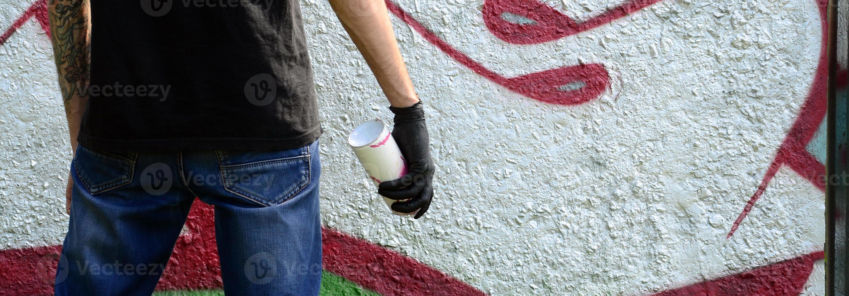 A young hooligan with a spray can stands against a concrete wall with graffiti paintings. Illegal vandalism concept. Street art photo