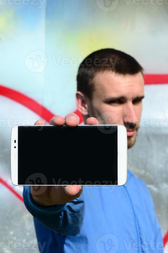 The graffiti artist demonstrates a smartphone with an empty black screen against the background of a colorful painted wall. Street art concept photo