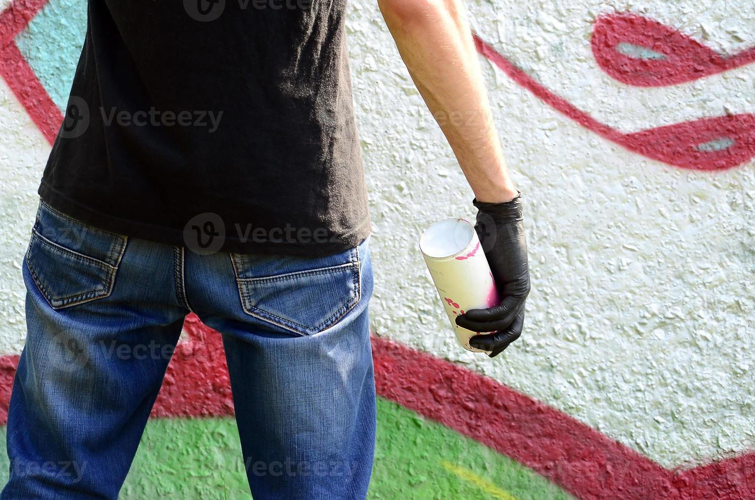 A young hooligan with a spray can stands against a concrete wall with graffiti paintings. Illegal vandalism concept. Street art photo