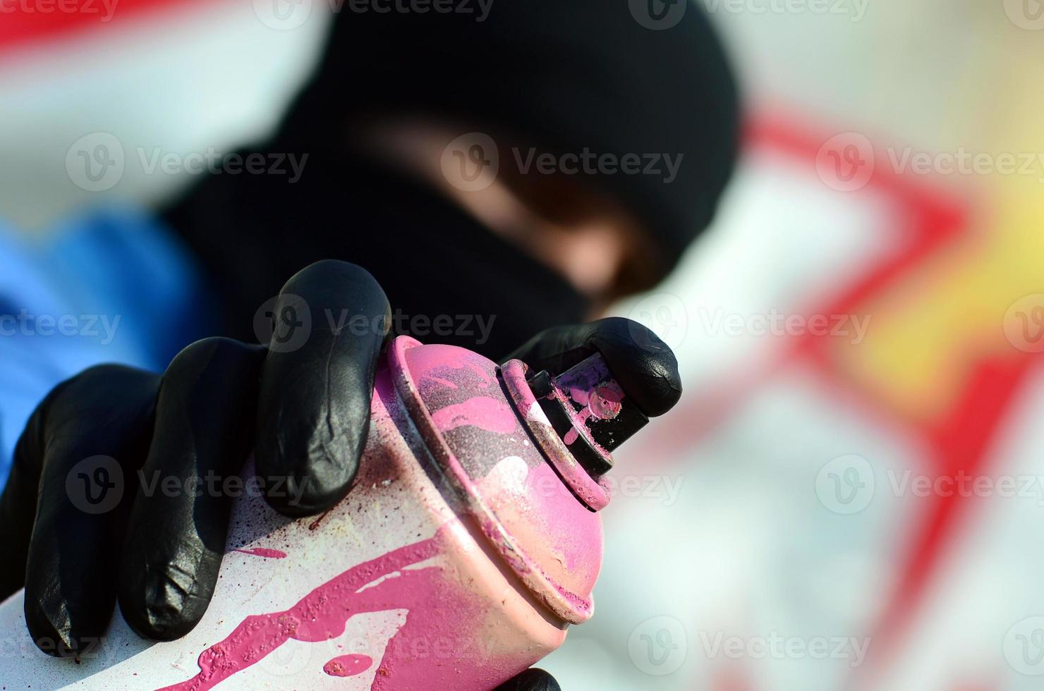 A young graffiti artist in a blue jacket and black mask is holding a can of paint in front of him against a background of colored graffiti drawing. Street art and vandalism concept photo