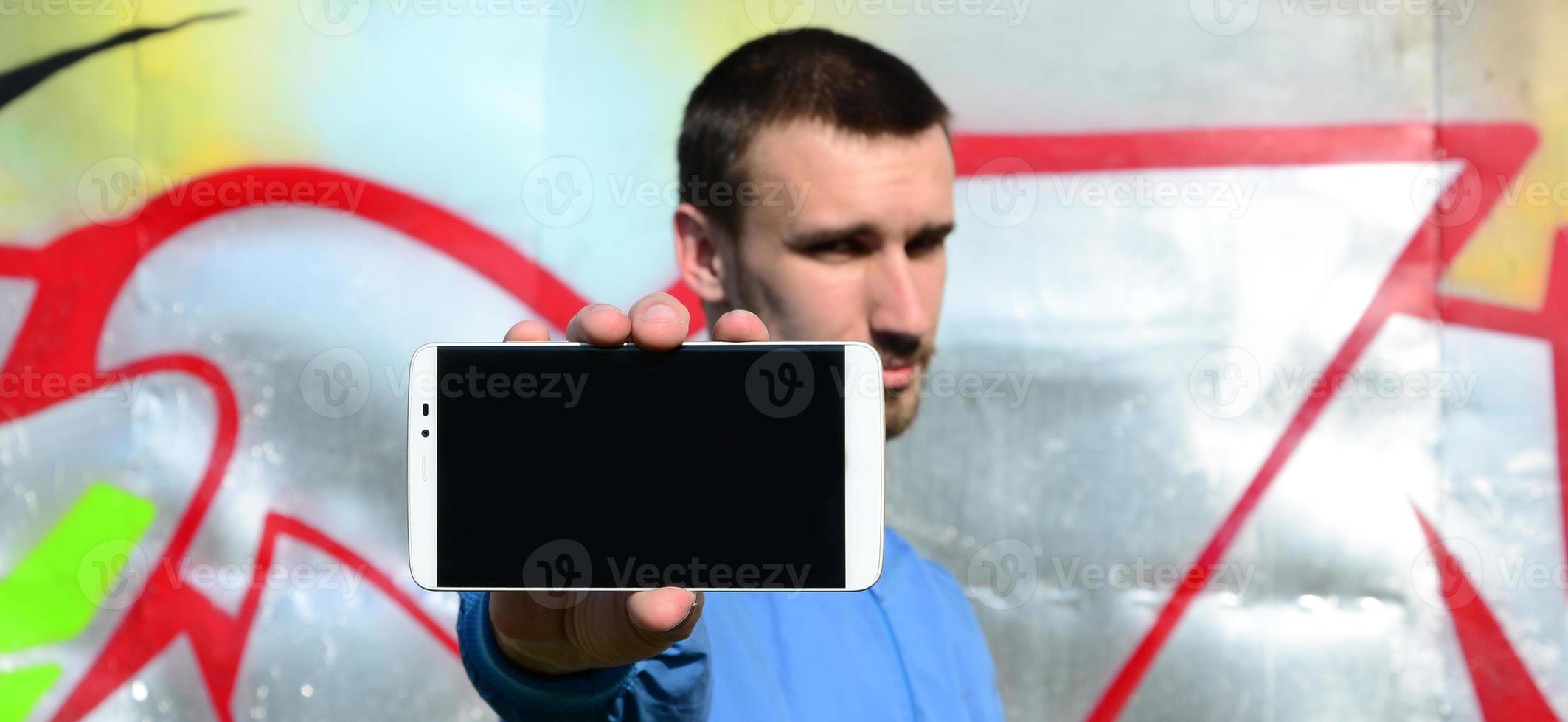 The graffiti artist demonstrates a smartphone with an empty black screen against the background of a colorful painted wall. Street art concept photo