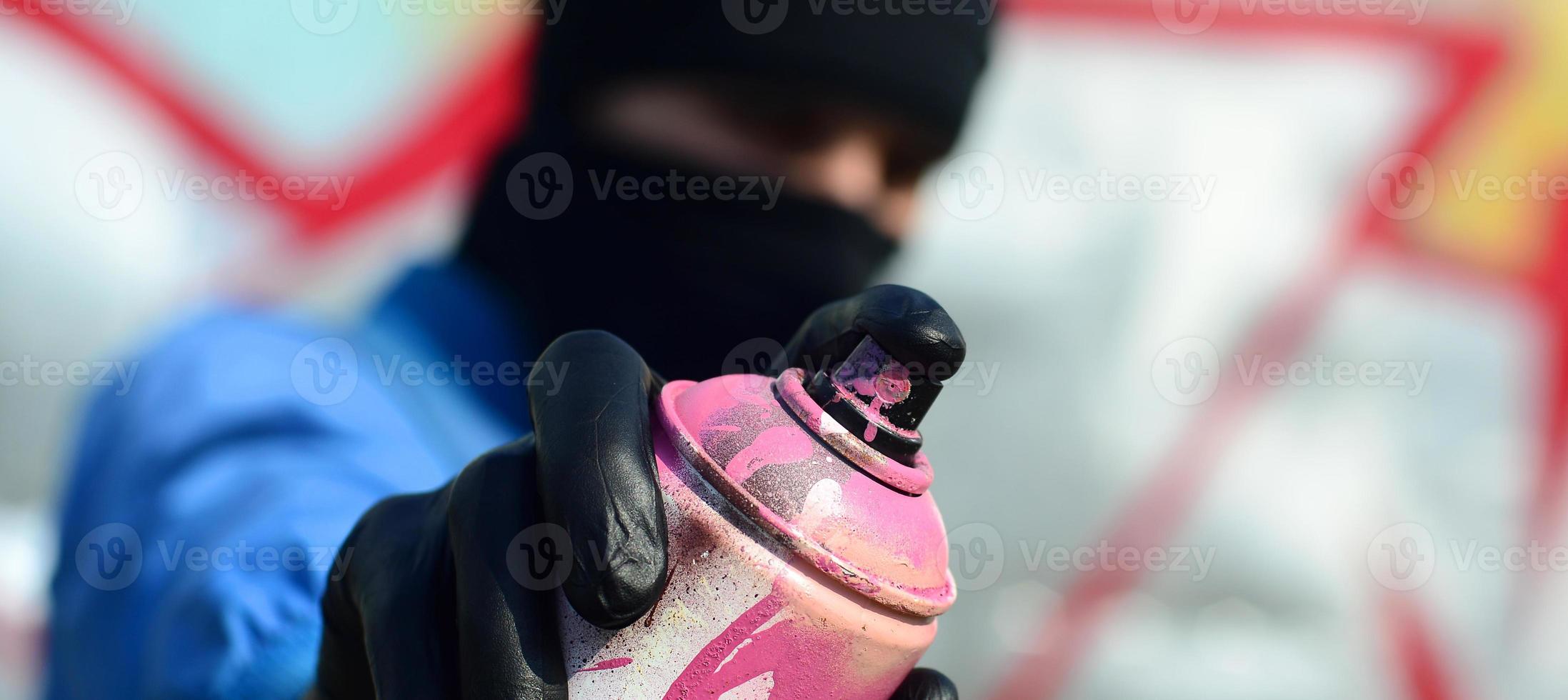 A young graffiti artist in a blue jacket and black mask is holding a can of paint in front of him against a background of colored graffiti drawing. Street art and vandalism concept photo