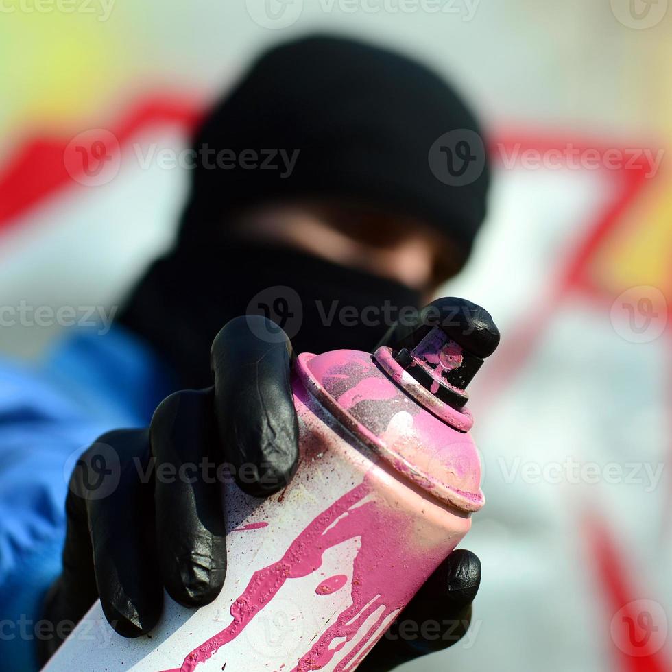 A young graffiti artist in a blue jacket and black mask is holding a can of paint in front of him against a background of colored graffiti drawing. Street art and vandalism concept photo