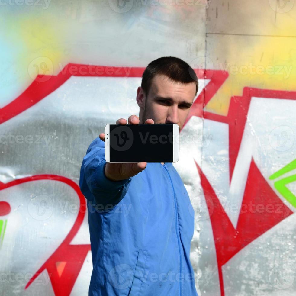 The graffiti artist demonstrates a smartphone with an empty black screen against the background of a colorful painted wall. Street art concept photo