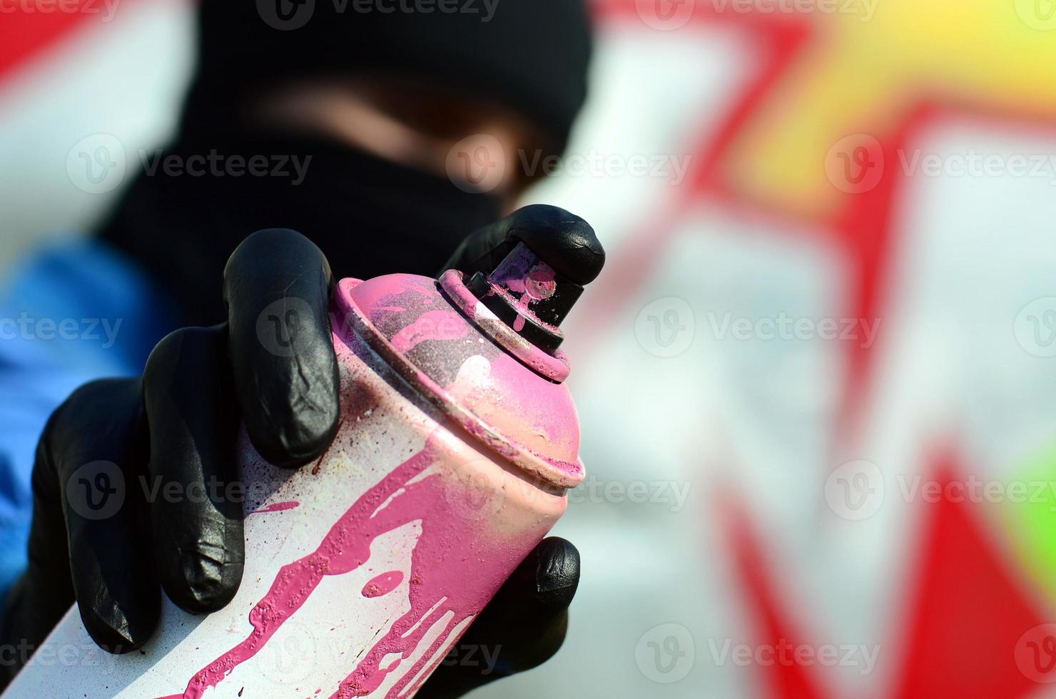 A young graffiti artist in a blue jacket and black mask is holding a can of paint in front of him against a background of colored graffiti drawing. Street art and vandalism concept photo
