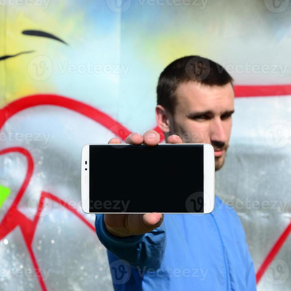 The graffiti artist demonstrates a smartphone with an empty black screen against the background of a colorful painted wall. Street art concept photo